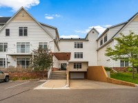 Front view of a multi-story white residential building on Old School Road, featuring an elevated entrance, stairs, ramp, and a small parking lot with vehicles in front of Unit 351 on the Mound. - Beise Real Estate