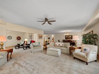 Spacious living room in Mound’s 2150 Old School Road, Unit 351, featuring beige carpet, sofas, armchairs, side tables with lamps, ceiling fan, and dining area in the background. Large window with curtains on the right. - Beise Real Estate