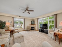 Spacious living room in Unit 351 at 2150 Old School Road, Mound, with beige carpet, multiple sofas, a ceiling fan, large windows, a cozy fireplace, and a TV on a stand. - Beise Real Estate