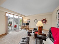 A living room in Unit 351 at 2150 Old School Road, Mound, features brown leather chairs, a lamp, wall art, a side table, and a desk by glass doors that open to a balcony with outdoor seating. - Beise Real Estate