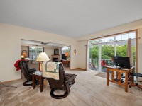 Living room in Unit 351 at 2150 Old School Road features a beige carpet, two recliners, a small desk with computer, and sliding glass doors opening to a balcony with peaceful views of trees. - Beise Real Estate