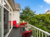 A small balcony at Unit 351, 2150 Old School Road, with black deck flooring, two black and red chairs, a red lounge chair, a round glass table with a potted plant, and a white railing, surrounded by green trees. - Beise Real Estate