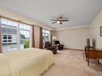 Spacious bedroom in Unit 351 with large windows, beige carpet, a bed with a yellow cover, two armchairs, a floor lamp, TV on a wooden stand, and framed art on a cream wall—steps from Mound and Old School Road. - Beise Real Estate