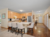 A modern kitchen at 2150 Old School Road, Unit 351, features light wood cabinets, stainless steel appliances, a center island with four upholstered bar stools, and an adjacent hallway leading to the front door in Mound. - Beise Real Estate