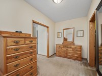 Unit 351 at 2150 Old School Road in Mound boasts a carpeted bedroom with two wooden dressers featuring brass handles, a wall mirror, framed artwork, and calming beige walls. - Beise Real Estate