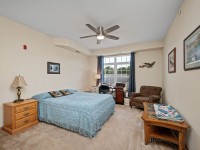 A bedroom at 2150 Old School Road, Unit 351, Mound, features a blue bedspread, wooden nightstand, lamp, armchair, coffee table, desk, large window with blue curtains, and walls adorned with paintings and decor. - Beise Real Estate