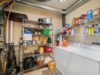 Laundry room in Unit 351, Old School Road, featuring a white washer and dryer, metal shelving with cleaning supplies, baskets, household items, and visible utility pipes along the ceiling and wall. - Beise Real Estate