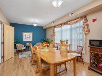 A dining room in Unit 351 on Old School Road features a wooden table and chairs, a sideboard with microwave, large window with vertical blinds, and a cozy sitting area with armchairs in the background. - Beise Real Estate
