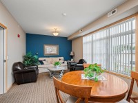 A living room in Unit 351 at 2150 Old School Road features a beige sofa, two dark armchairs, a round coffee table, and a dining table with chairs by a large window with vertical blinds. - Beise Real Estate
