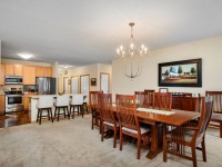 Open dining and kitchen area at Unit 351, Old School Road, with wooden dining table, chairs, bar stools at the counter, chandelier, and a wall-mounted landscape painting. Kitchen features stainless steel appliances. - Beise Real Estate
