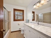 Bathroom at 4709 Utah Avenue N features a marble countertop, white vanity, toilet, glass shower enclosure, large mirror, and a window with wooden trim—offering comfort and style in the heart of New Hope. - Beise Real Estate