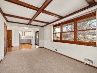 A bright living room on 4709 Utah Avenue features carpeted flooring, wood ceiling beams, large windows, and an open view into a kitchen with hardwood floors and white cabinets in the heart of New Hope. - Beise Real Estate