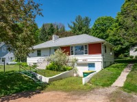 Charming single-story white house with a gray roof, red accents, and a front ramp at 4880 Grand View Avenue, Minnetrista—surrounded by trees and a lush green lawn on a sunny day. - Beise Real Estate