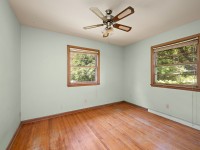 Bright, empty room on Grand View Avenue in Minnetrista featuring light green walls, two wooden-framed windows, a ceiling fan, and worn hardwood flooring. - Beise Real Estate