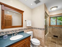 Bathroom with wood vanity, dark countertop, large mirror, toilet, beige tile walls and floor, towel rack, walk-in shower, and window with view of greenery. - Beise Real Estate