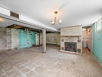 Unfinished basement at 4880 Grand View Avenue, Minnetrista, featuring exposed pipes, turquoise cinder block walls, tile floor, a white brick fireplace, and ceiling beams; minimal lighting and visible wear throughout—a unique real estate opportunity. - Beise Real Estate