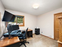 Home office with three monitors on two wooden desks, a black office chair, filing cabinet, wooden cabinet, and window with outdoor view. Beige walls and carpet flooring. - Beise Real Estate