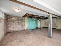 Unfinished basement at 4880 Grand View Avenue, Minnetrista, featuring exposed ceiling pipes, damaged walls, cinder blocks, and a tiled floor. Real estate highlights include ceiling light and some peeling paint visible. - Beise Real Estate