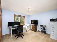A home office with a desk, dual monitors, office chair, filing cabinets, TV, printer, and camera equipment on beige carpet with light blue walls and a window. - Beise Real Estate