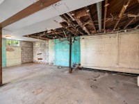 Unfinished basement at 4880 Grand View Avenue, Minnetrista, with exposed pipes and beams, damaged ceiling, painted cinderblock walls, and a concrete floor. Natural light enters through a small window. - Beise Real Estate