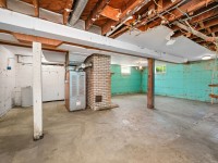 Unfinished basement at 4880 Grand View Avenue, Minnetrista, with exposed beams, pipes, and ductwork, concrete floor, brick chimney, and a teal painted cinder block wall. - Beise Real Estate