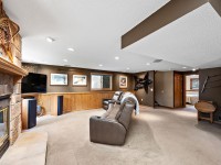 Basement living room with a stone fireplace, leather recliners facing a TV, carpeted floor, wood paneling, and rustic decor on the walls. - Beise Real Estate