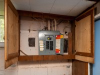 An open wooden cabinet at 4880 Grand View Avenue reveals an electrical breaker panel mounted on a worn basement wall with exposed wiring and peeling paint. - Beise Real Estate