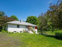 Charming single-story white house with a red door at 4880 Grand View Avenue, Minnetrista, surrounded by green grass, trees, and a clear blue sky on a sunny day. - Beise Real Estate
