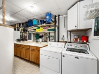 Laundry room with washer, dryer, utility sink, wooden cabinets, shelves with storage bins and towels, and a water heater on the left. Clothes hangers and cleaning supplies are visible. - Beise Real Estate