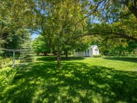 A small white house sits in a grassy yard surrounded by large green trees under a clear blue sky at 4880 Grand View Avenue, Minnetrista. - Beise Real Estate