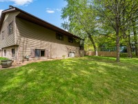 A beige, split-level house with brown trim sits on a well-maintained lawn with mature trees and a wooden fence in the backyard. - Beise Real Estate