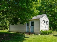 Charming small white building with two blue doors and a single exterior light, nestled among green trees and grass on a sunny day along Grand View Avenue in Minnetrista. - Beise Real Estate