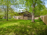 Single-story house with a raised deck, surrounded by a green lawn and mature trees, next to a wooden fence on a sunny day. - Beise Real Estate