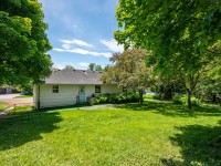 A single-story house with light-colored siding, a shingled roof, and a blue door sits on 4880 Grand View Avenue in Minnetrista, surrounded by a grassy yard and mature green trees on a sunny day. - Beise Real Estate