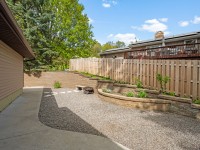 Backyard with gravel ground, a fire pit, curved retaining walls with plants, wooden privacy fence, and concrete walkway beside a house. - Beise Real Estate
