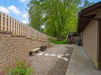 Side yard with gravel path, stone bench, retaining wall, wooden fence, plants, and stairs leading to house with beige siding. - Beise Real Estate