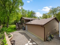 Rear view of a tan house with a wooden deck, stairs, gravel area, hose reels, trash bins, and a fenced backyard with green trees and lawn. - Beise Real Estate