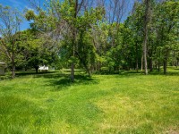 A grassy yard with scattered trees and two small white buildings partially visible in the background under a blue sky at 4880 Grand View Avenue, Minnetrista. - Beise Real Estate