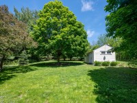 A small white shed sits next to a large leafy tree in the grassy yard at 4880 Grand View, surrounded by dense green foliage under a bright blue sky. - Beise Real Estate