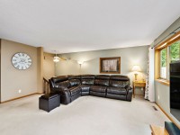 A living room with a large black leather sectional sofa, beige carpet, a wall clock, lamps, a framed picture, and a window with a view of trees. - Beise Real Estate