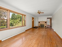 A long living room with wood floors, a ceiling fan, brick fireplace, and large windows overlooking a green yard. Walls are white and the room is unfurnished—perfect for your vision at 4880 Grand View Avenue in Minnetrista real estate. - Beise Real Estate