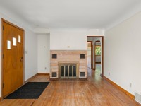 Living room with wood floors, a wooden front door on the left, a brick fireplace in the center, and a doorway leading to another room with visible greenery outside. Located in Minnetrista real estate at 4880 Grand View Avenue. - Beise Real Estate