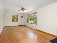 Bright and inviting, this Minnetrista real estate gem at 4880 Grand View Avenue features an empty living room with hardwood floors, white walls, a ceiling fan, AC unit, wooden door, and natural light streaming through two windows. - Beise Real Estate