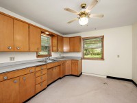 A kitchen at 4880 Grand View Avenue features wooden cabinets, a ceiling fan with a light, two windows, a countertop, and a beige floor. No appliances are visible in this Minnetrista home. - Beise Real Estate