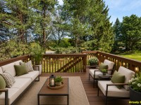 Virtually staged image of a wooden deck with outdoor seating, cushions, potted plants, and a coffee table, overlooking a green yard and trees in scenic Eden Prairie on Wiedman Way. - Beise Real Estate