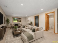 A living room at Wiedman Way, Eden Prairie, with a beige sectional sofa, coffee table, TV, dining set, large window, and an open door to a bathroom. Room is labeled as virtually staged. - Beise Real Estate