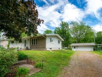 Charming single-story house on Rainbow Avenue in Maple Plain, featuring a front porch, green lawn, trees, and a gravel driveway leading to a detached garage under a partly cloudy sky. - Beise Real Estate