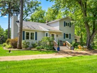 Charming two-story light gray house on Old Crystal Bay Road in Orono, featuring a front porch, lush green grass, mature trees, and inviting outdoor seating—perfect for sunny days. - Beise Real Estate