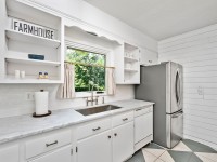 Bright kitchen on Old Crystal Bay Road with white cabinets, marble countertops, stainless steel refrigerator, farmhouse décor, and a window with curtains above the sink. - Beise Real Estate