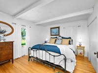 A bright Orono bedroom on Old Crystal Bay Road features a metal bed frame, white bedding with blue accents, a wood dresser, hardwood floor, and windows with white curtains letting in natural light. - Beise Real Estate